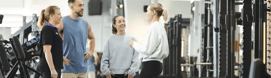 A group of four individuals converses in a gym, surrounded by exercise equipment, discussing fitness routines and training.