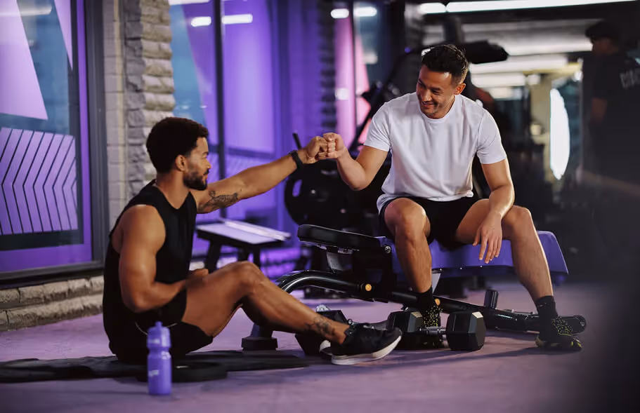 Two young men in a gym exchange a fist bump, celebrating fitness and camaraderie, with weights and workout equipment around them.